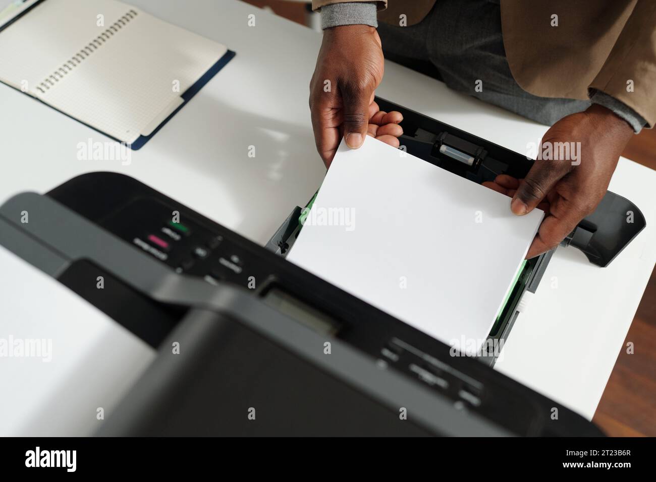 Au-dessus de l'angle des mains du jeune homme afro-américain employé de bureau mettant du papier dans le bac de la machine xerox debout sur le bureau avec bloc-notes Banque D'Images