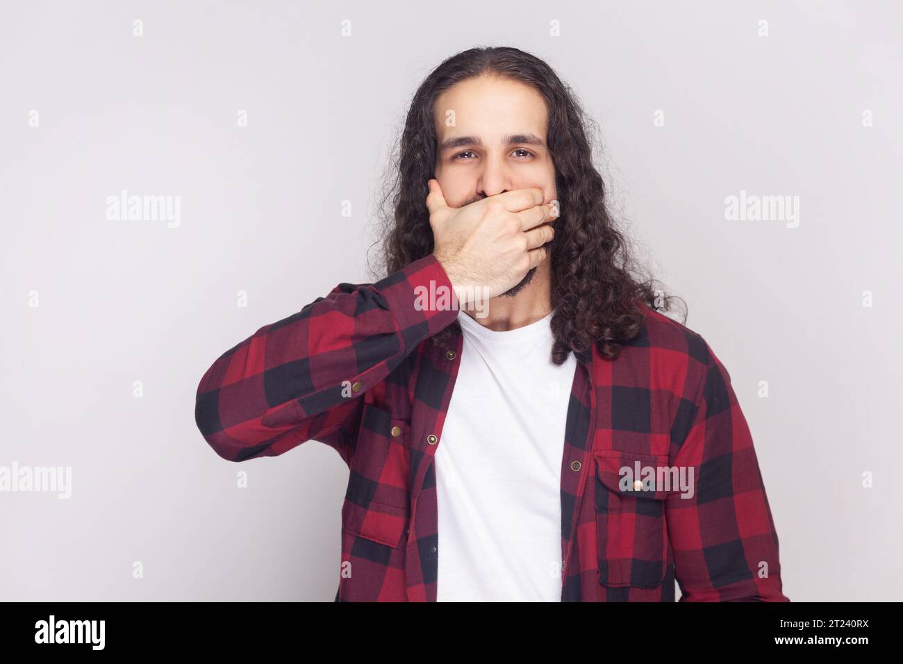Portrait d'homme barbu avec de longs cheveux bouclés en chemise rouge à carreaux essaie de ne pas dire secret à ses amis, couvre la bouche tout en commérant. Studio intérieur tourné isolé sur fond gris. Banque D'Images