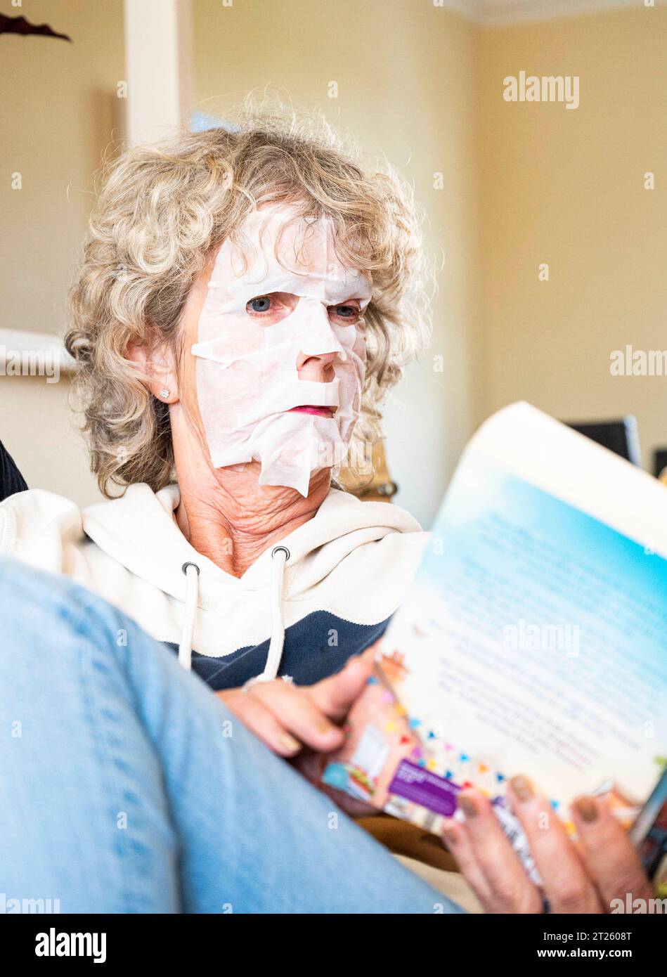 Femme mature lisant un livre à la maison portant un pack visage pour essayer de se débarrasser des rides indésirables et garder sa peau lisse Banque D'Images