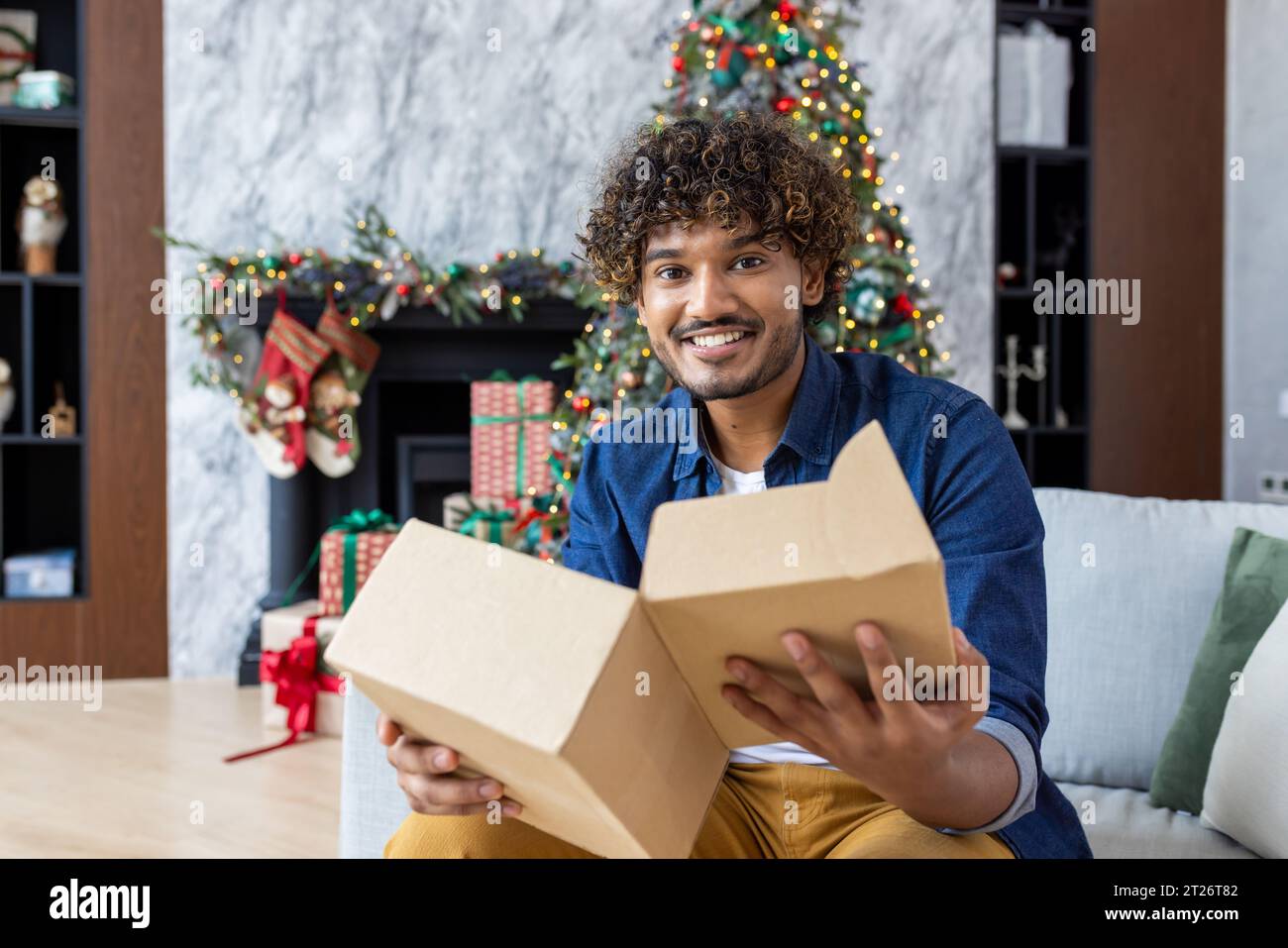 Portrait de l'homme heureux à la maison à Noël, homme hispanique tenant le cadeau de boîte aux lettres en carton, heureux reçu le produit de la boutique en ligne à l'heure, assis dans le salon près de l'arbre de Noël. Fête du nouvel an. Banque D'Images