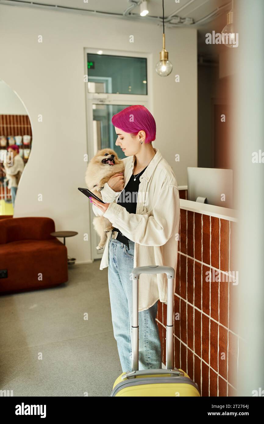 femme élégante avec spitz pomeranian et smartphone près de la valise et du bureau de réception dans l'hôtel pour animaux de compagnie Banque D'Images