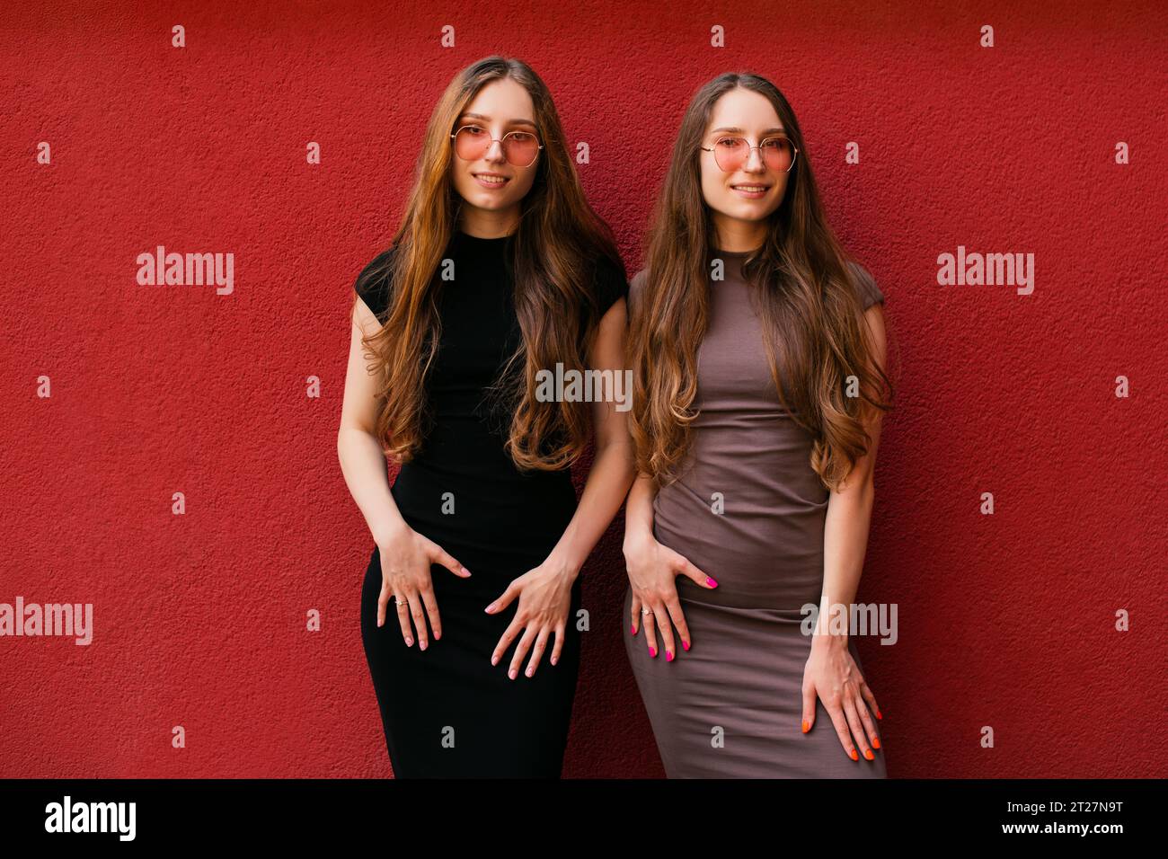 Mignon deux jeunes sœurs jumelles décontractées drôles avec des lunettes de soleil à la mode regardant la caméra debout sur un fond de mur rouge en plein air jour d'été Banque D'Images