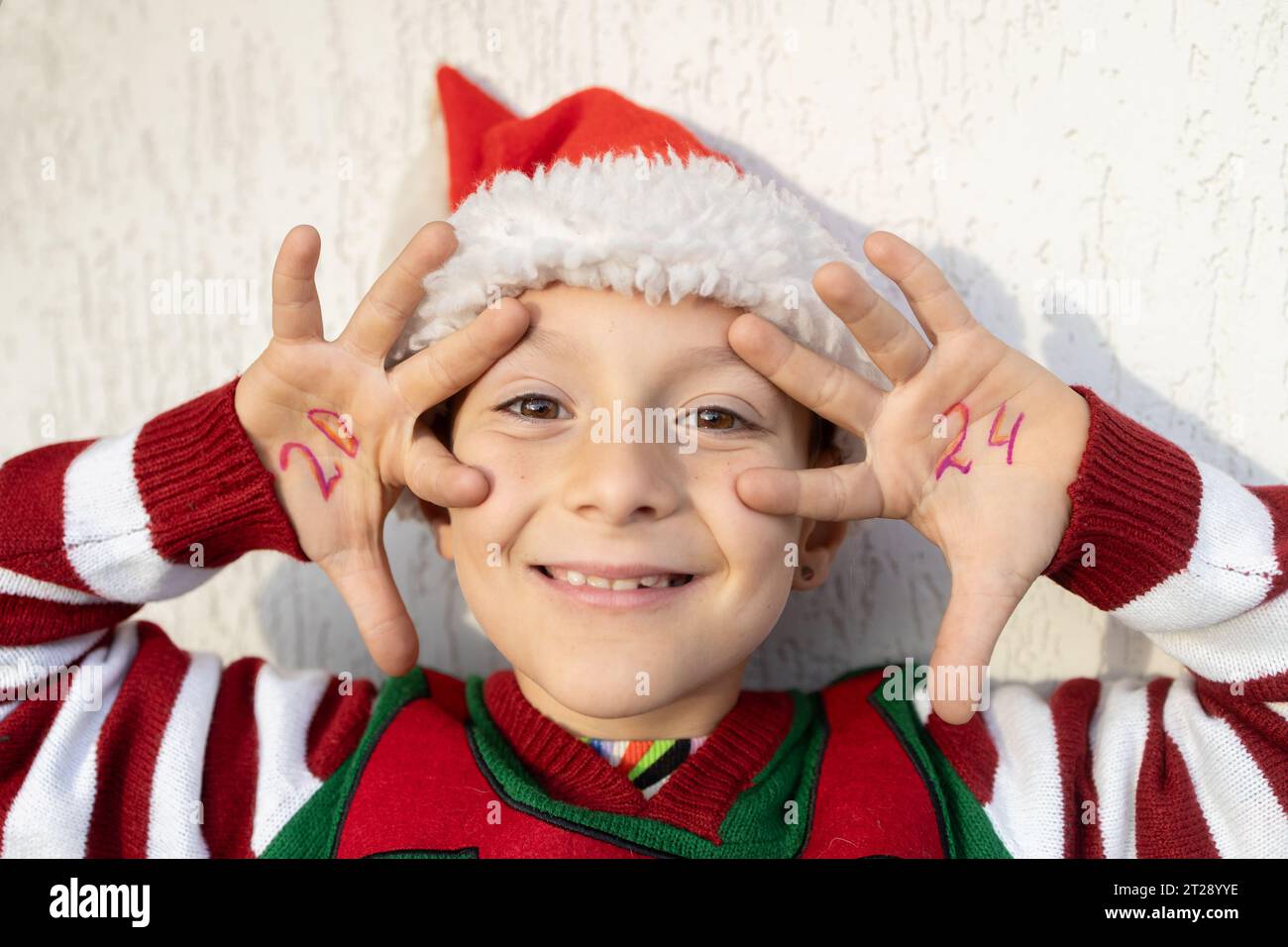 Portrait de visage de souriant joyeux enfant mignon dans le chapeau de Père Noël et pull elfe, numéros de la nouvelle année 2024 Banque D'Images