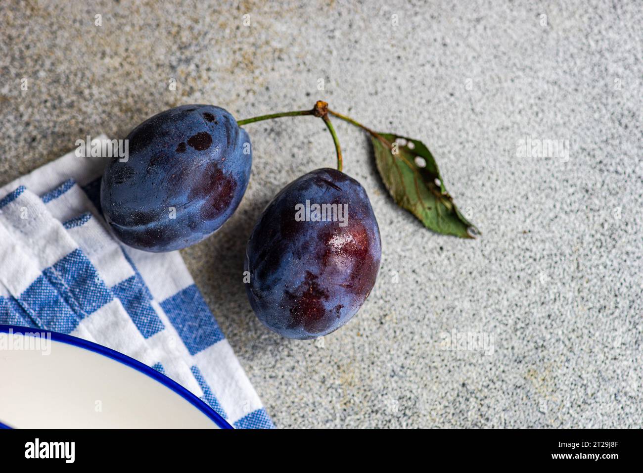 Gros plan de prunes mûres placées sur une surface grise près d'un bol en céramique et d'une serviette rayée Banque D'Images