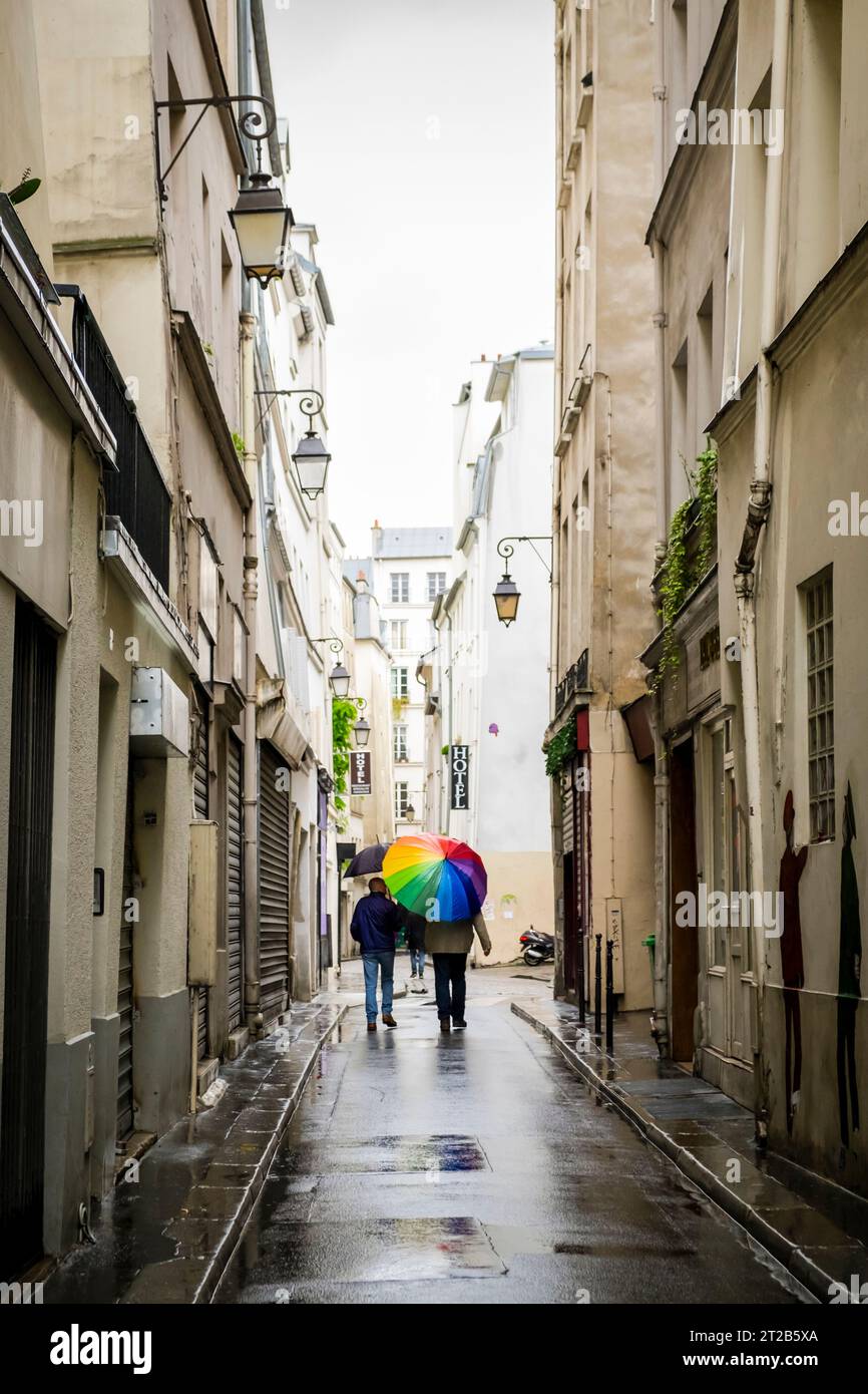 Les gens marchent le long d'une rue étroite trempée par la pluie avec un parapluie de couleur arc-en-ciel dans le quartier du Marais, Paris, France. Banque D'Images