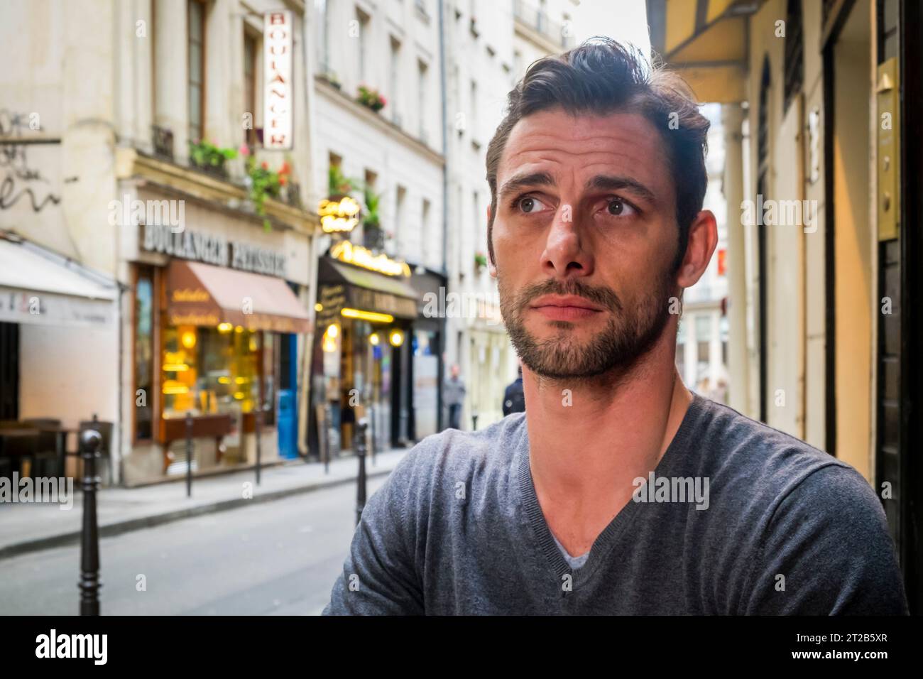 Bel homme regarde loin de la caméra avec un regard sérieux sur son visage tout en étant assis dans un café en plein air à Paris, France. Banque D'Images