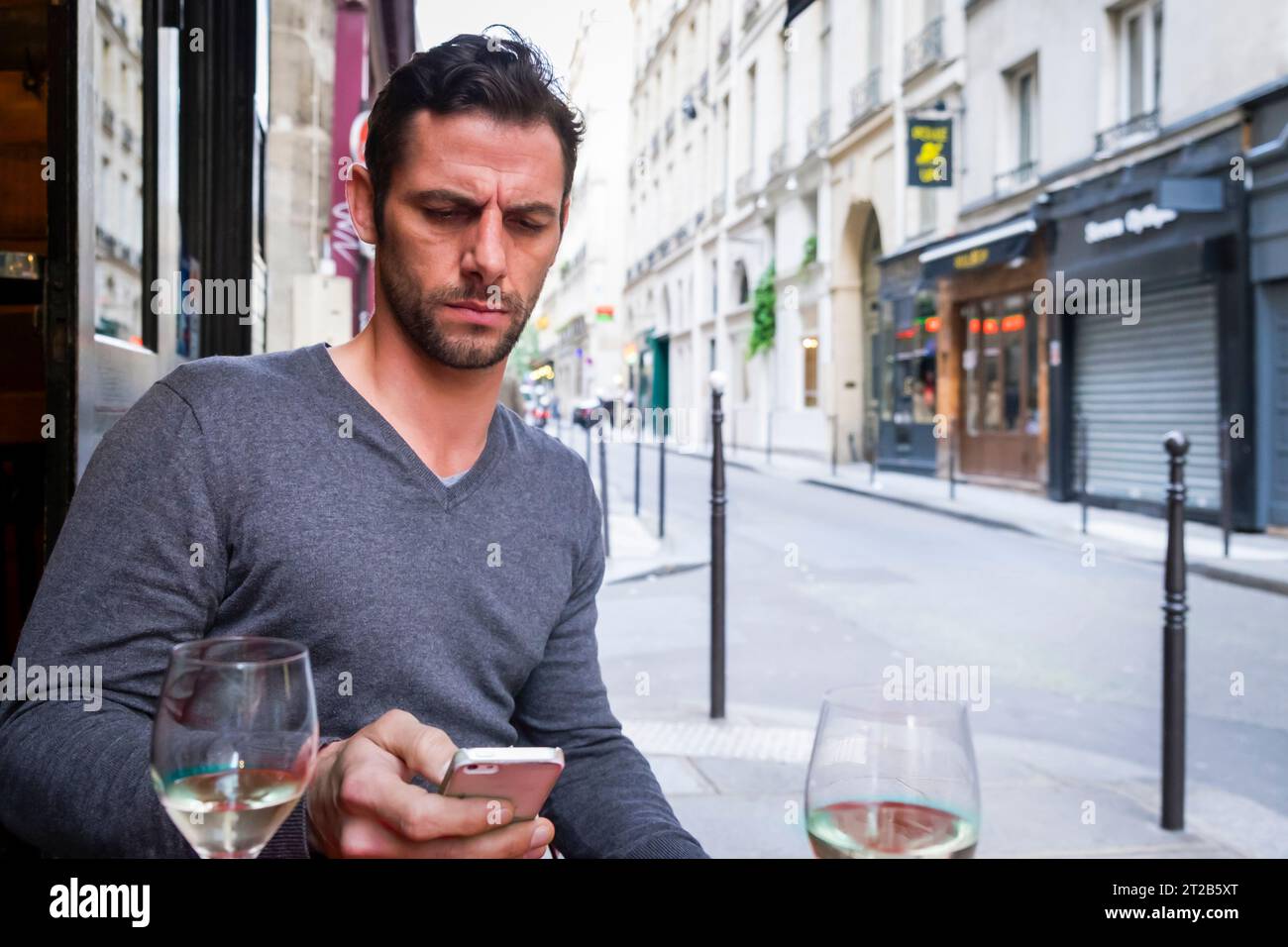 Bel homme regarde un téléphone portable et fronce les sourcils tout en étant assis dans un restaurant en plein air à Paris, France. Banque D'Images