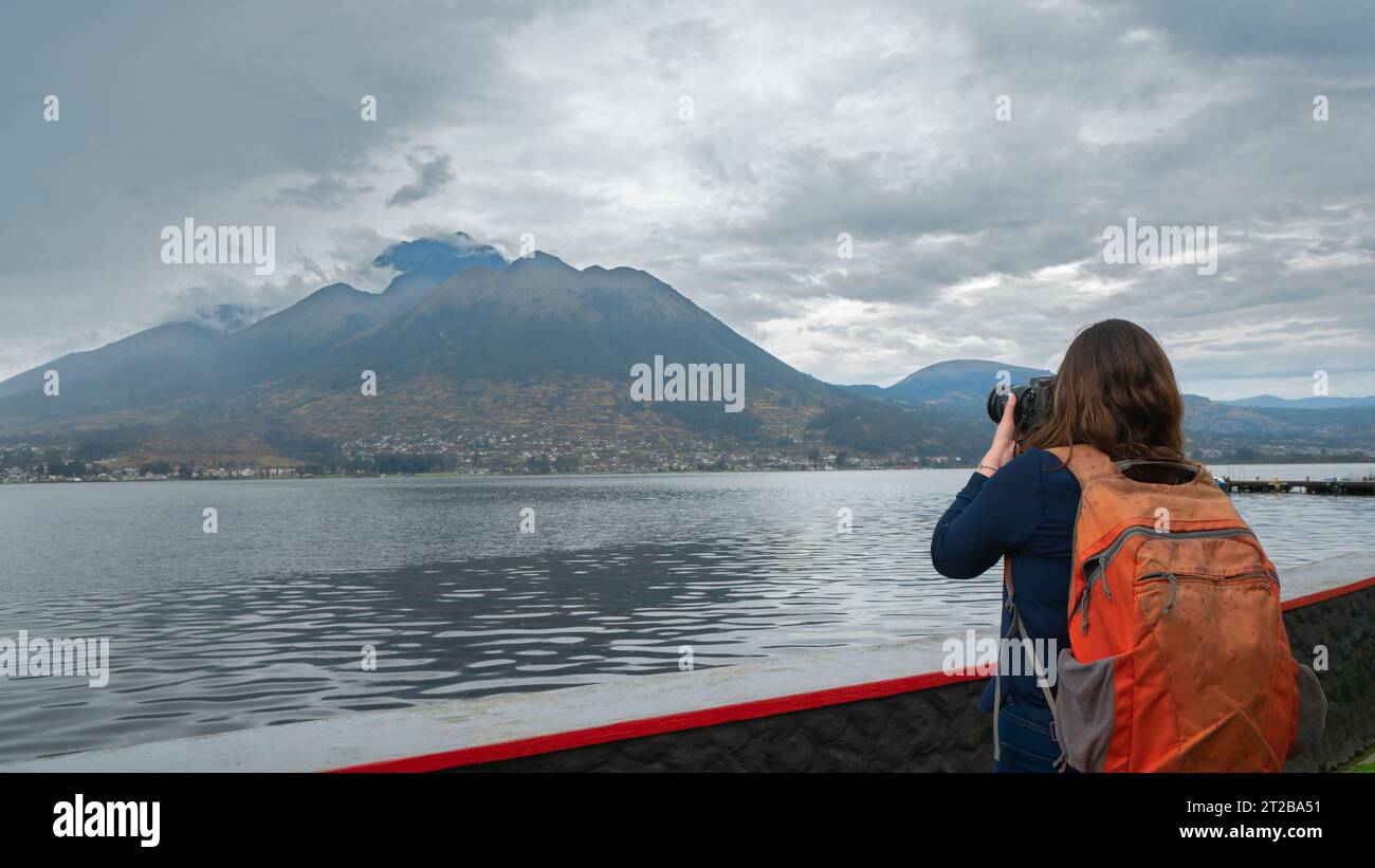 Belle jeune femme latino-américaine vêtue de bleu avec sac à dos orange vue de derrière en prenant une photo du volcan Imbabura depuis le lac San Pablo Banque D'Images