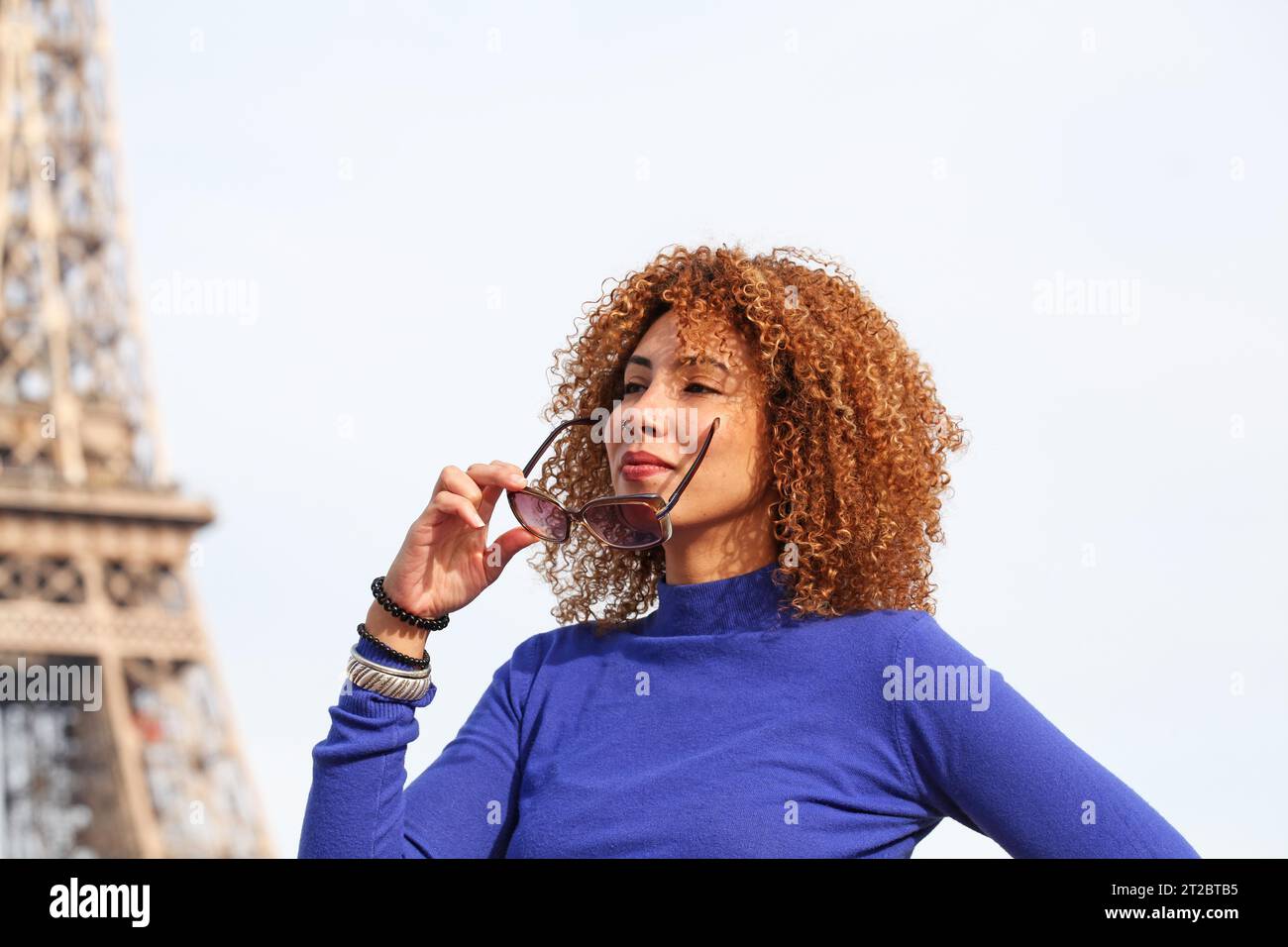 JOYEUX CHEVEUX BOUCLÉS JEUNE FEMME EN PULL BLEU TENANT SES LUNETTES DE SOLEIL À LA TOUR EIFFEL Banque D'Images
