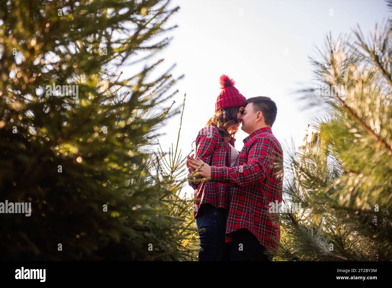 Joyeux couple amoureux dans des chemises rouges à carreaux, chapeaux tricotés se trompent, riant parmi le marché vert de l'arbre de Noël. Le jeune homme tient les mains de Banque D'Images