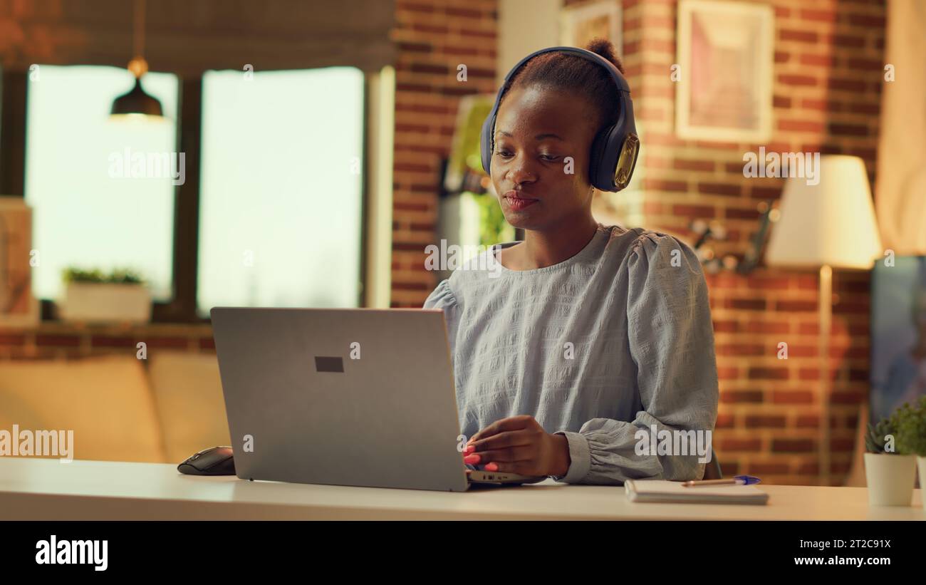 Télétravailleur écoutant de la musique et résolvant des tâches quotidiennes, femme indépendante. Fille afro-américaine s'amusant avec casque pendant qu'elle travaille sur des tâches pendant l'heure de coucher du soleil à la maison. Prise de vue à main levée. Banque D'Images
