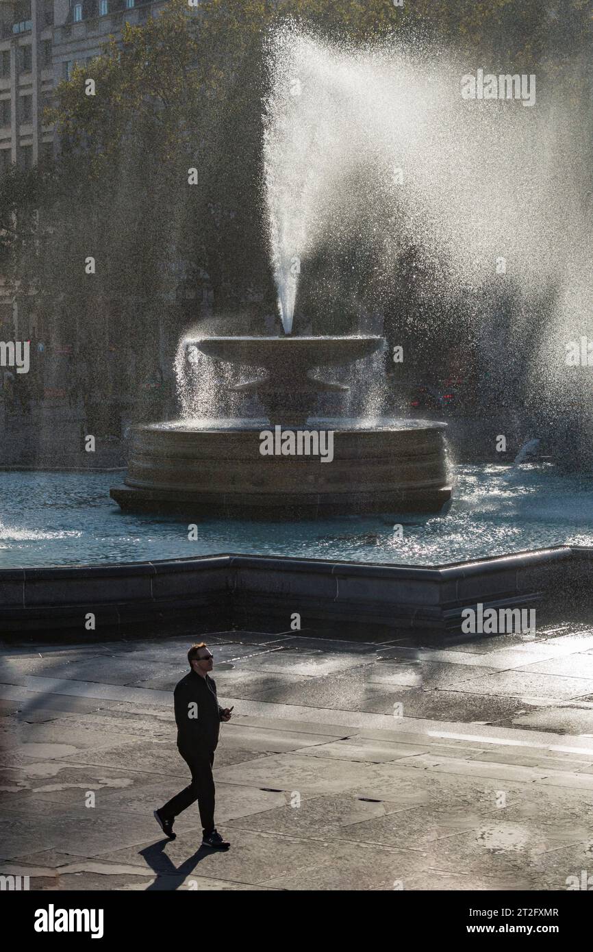 Homme marchant devant l'une des fontaines de Trafalgar Square, Londres, un jour d'automne venteux Banque D'Images