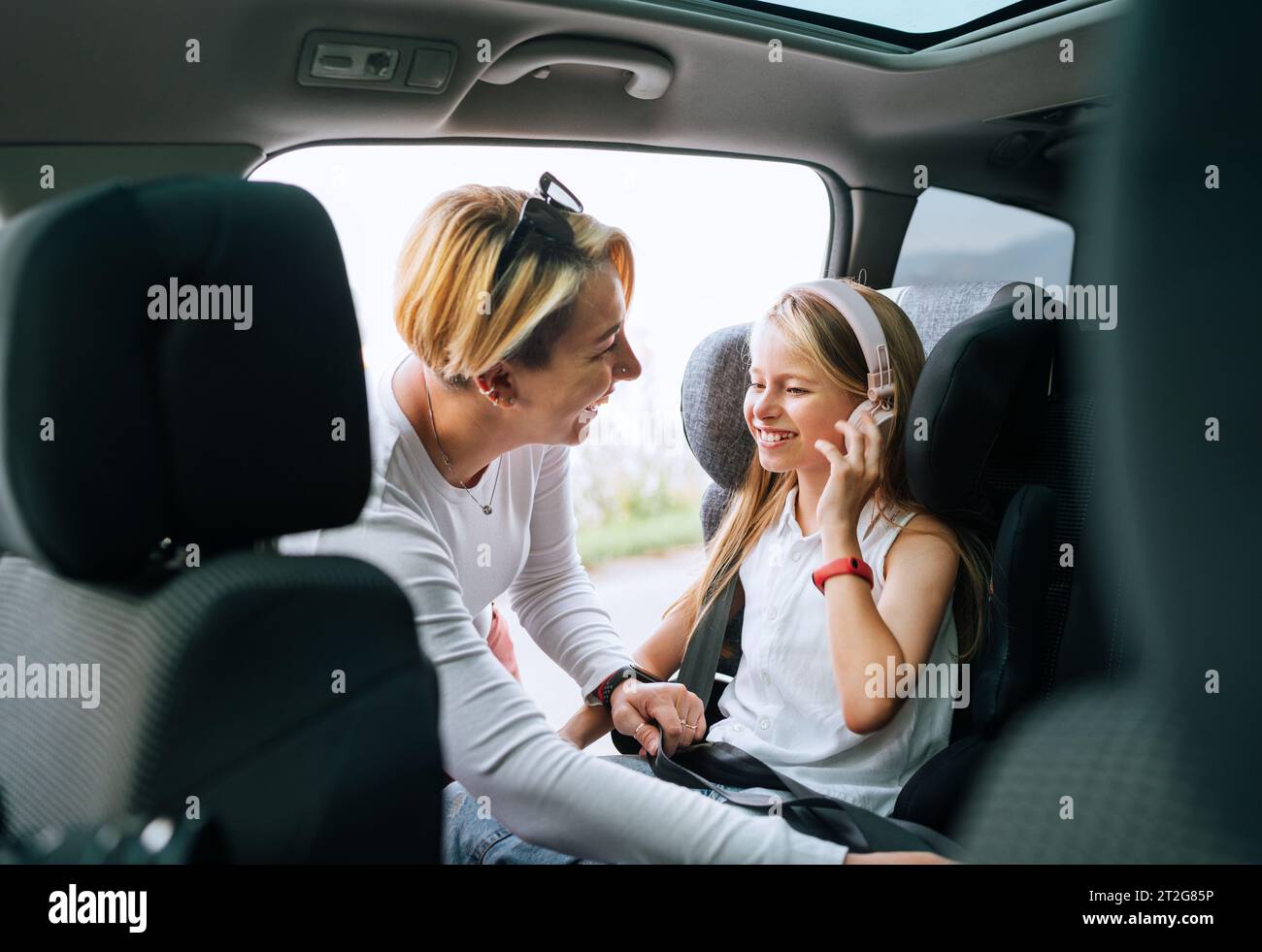 À l'intérieur de la photo de voiture d'une mère fixant avec la ceinture de sécurité automatique sa petite fille assise dans le siège d'enfant. Fille écoutant de la musique à l'aide d'écouteurs Banque D'Images