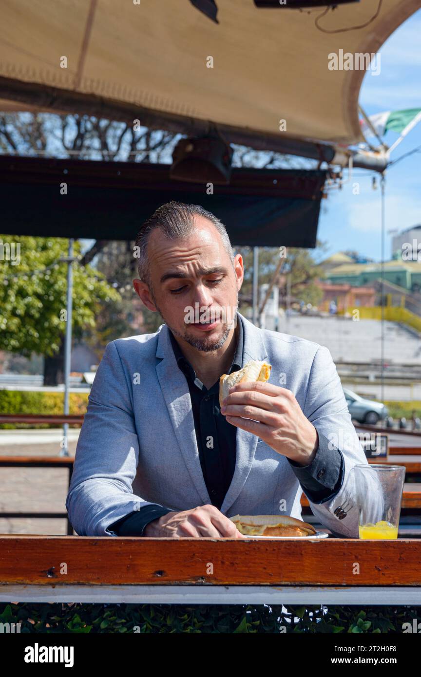 Image verticale d'un homme caucasien adulte, assis à l'extérieur du restaurant, profitant d'une journée heureuse voyant à quel point il mange un sandwich délicieux. espace de copie. Banque D'Images