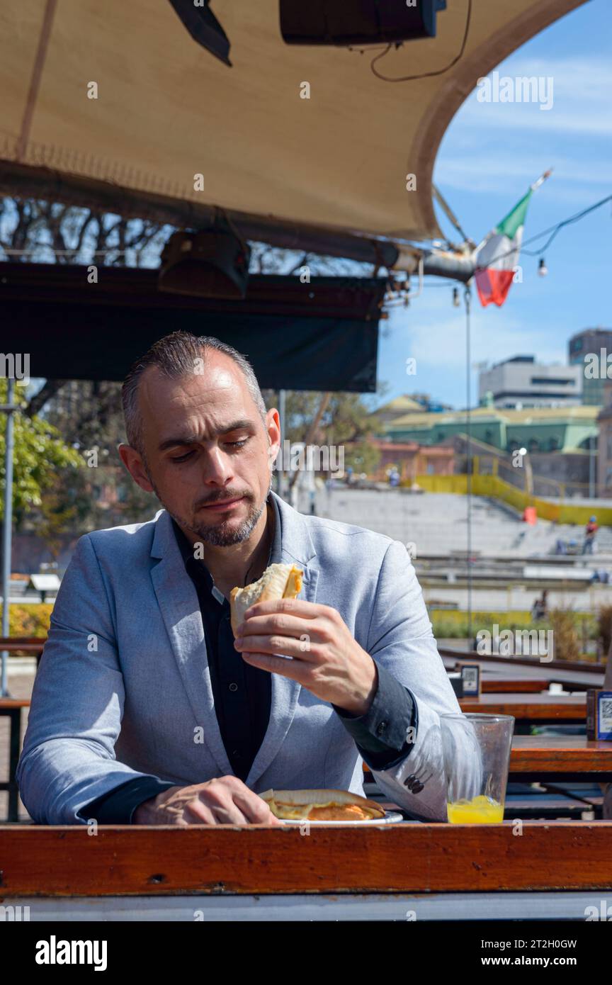 Image verticale d'un homme caucasien adulte, assis à l'extérieur du restaurant, profitant d'une journée heureuse voyant à quel point il mange un sandwich délicieux. espace de copie. Banque D'Images