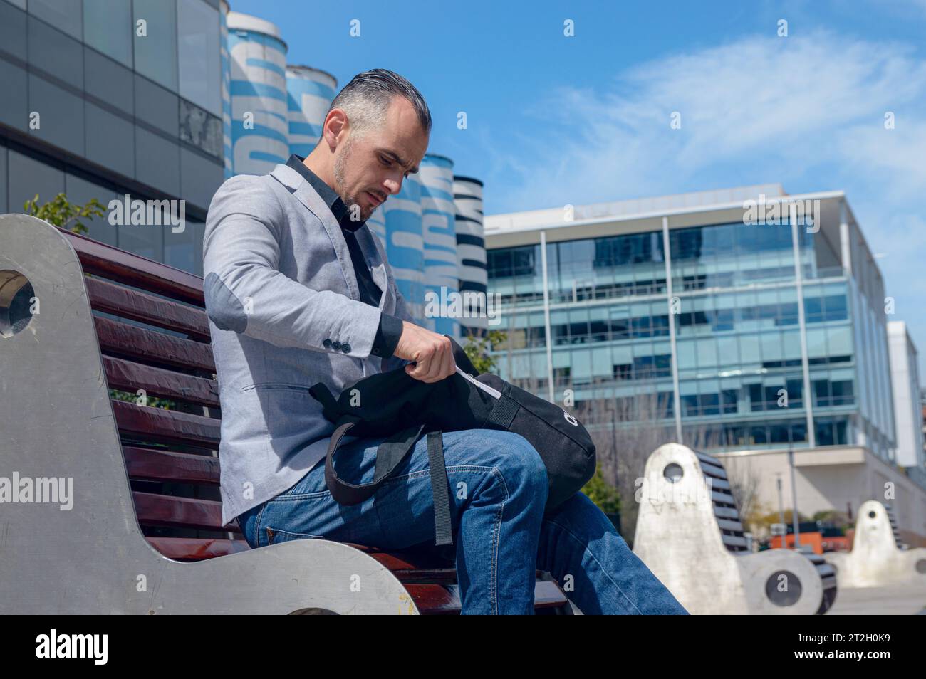 Homme d'affaires adulte caucasien, assis à l'extérieur des bâtiments de travail à la recherche de quelque chose dans son sac à dos, portant une veste élégante et un pantalon en jean, peo d'affaires Banque D'Images