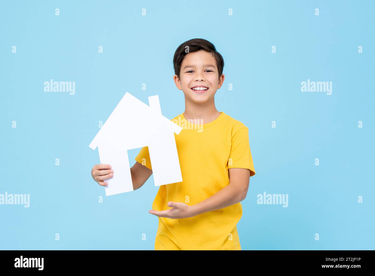 Garçon de race mixte optimiste dans le t-shirt jaune souriant et montrant la maison de papier pendant la campagne de promotion de l'agence immobilière sur fond bleu Banque D'Images