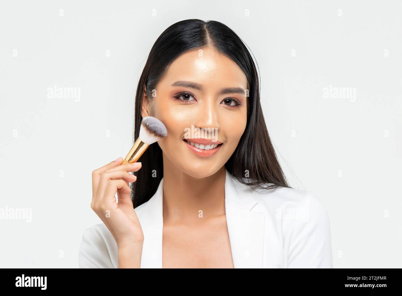 Femme asiatique joyeuse regardant la caméra avec le sourire et appliquant du blush sur la joue pendant la routine de beauté sur fond blanc Banque D'Images
