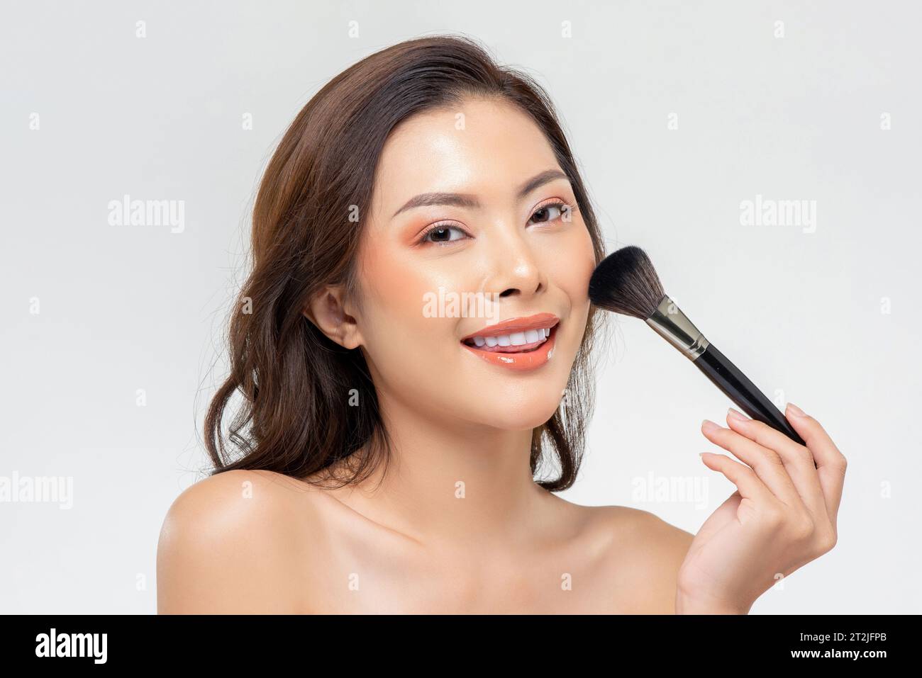 Modèle féminin asiatique positif regardant la caméra avec le sourire tout en appliquant le maquillage avec un pinceau doux sur fond blanc Banque D'Images