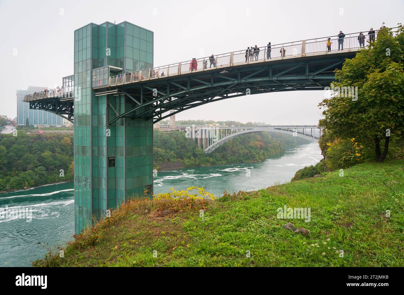 Les visiteurs bordent la promenade de la tour d'observation en verre vert au-dessus de la gorge, avec le Rainbow Bridge au loin, au parc d'État de Niagara Falls, New York. Banque D'Images
