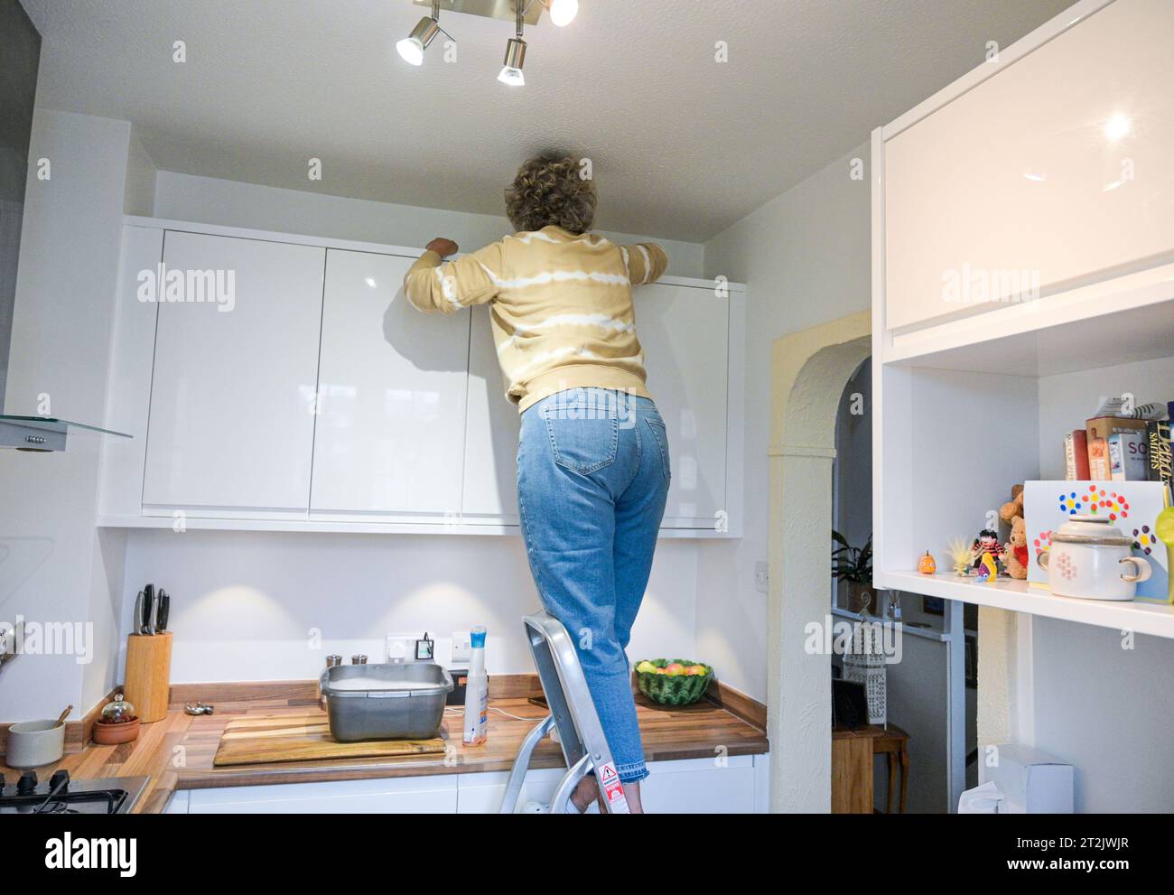 Femme d'âge moyen sur un escabeau nettoyant ses placards de cuisine avec un bol d'eau savonneuse Banque D'Images