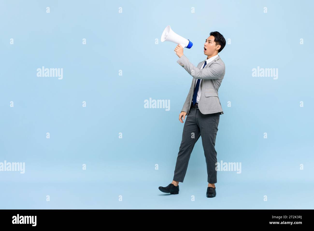 Jeune bel homme asiatique en costume d'affaires formel tenant mégaphone dans fond de studio de couleur bleu clair isolé avec espace de copie Banque D'Images