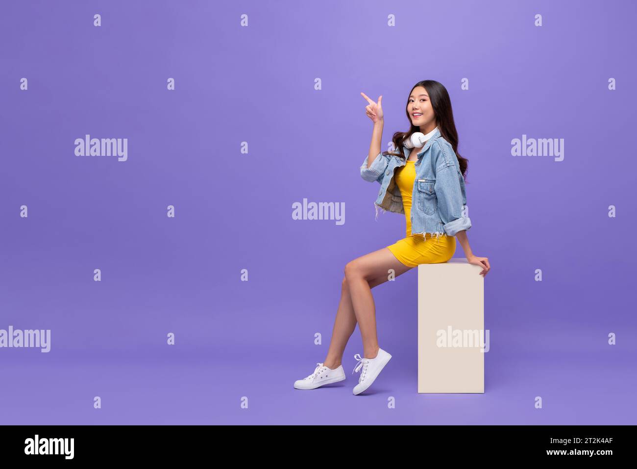 Jolie femme asiatique souriante assise et pointant du doigt vers le haut dans fond de studio de couleur violet isolé avec espace de copie Banque D'Images