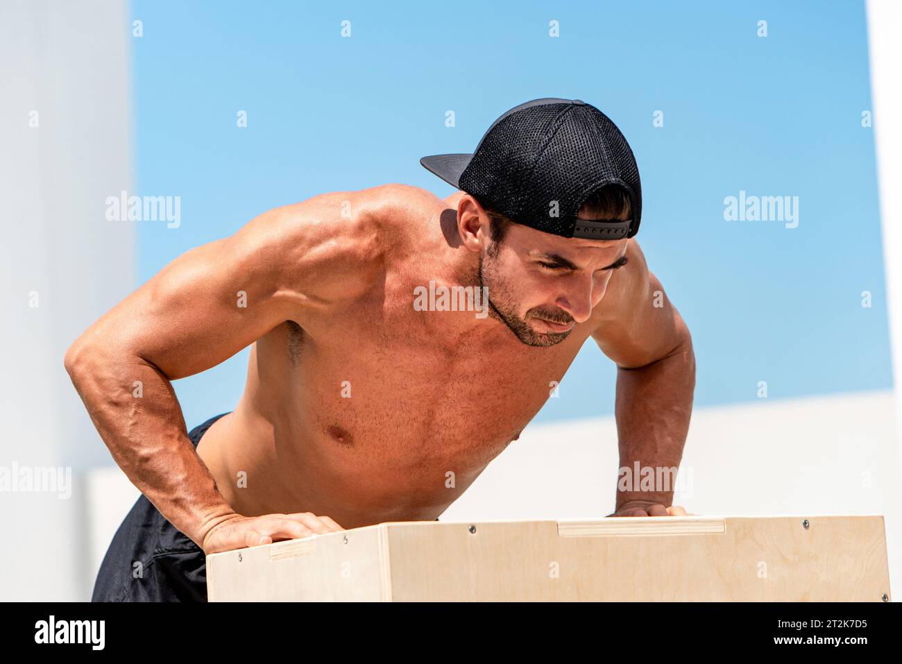 Athlète hispanique masculin dévoué sans chemise portant une casquette et faisant des pompes sur la boîte pliométrique en plein air contre le ciel d'été Banque D'Images