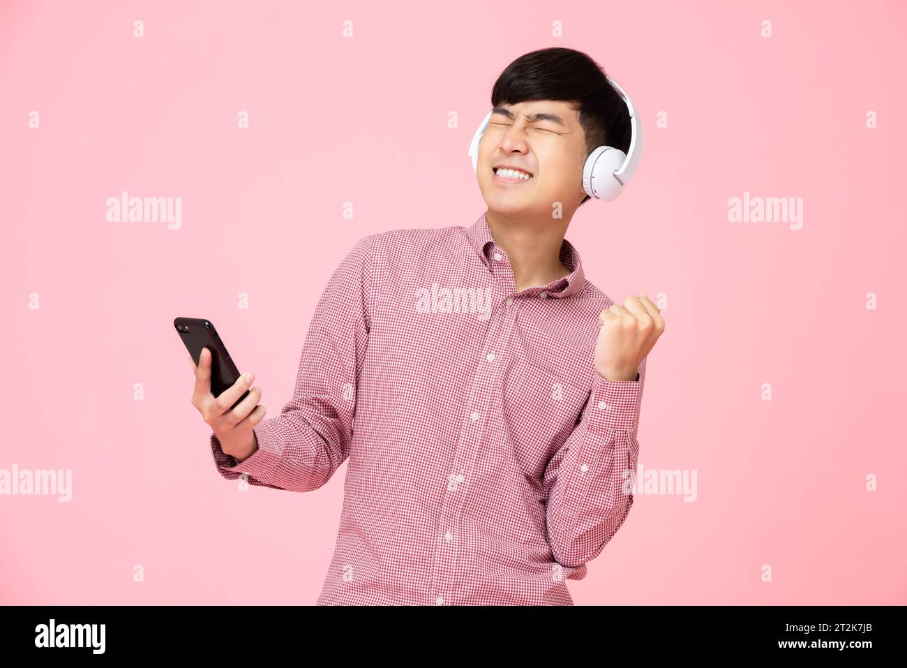 Homme asiatique extatique portant des écouteurs écoutant de la musique en ligne en streaming à partir d'un téléphone portable avec son poing vers le haut dans le fond rose studio Banque D'Images