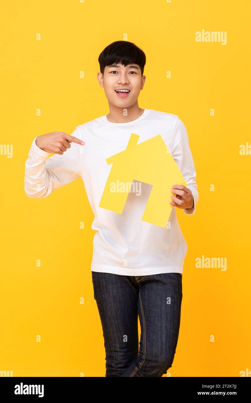 Souriant bel homme asiatique tenant et pointant la main à la maison modèle sur fond isolé de studio jaune Banque D'Images