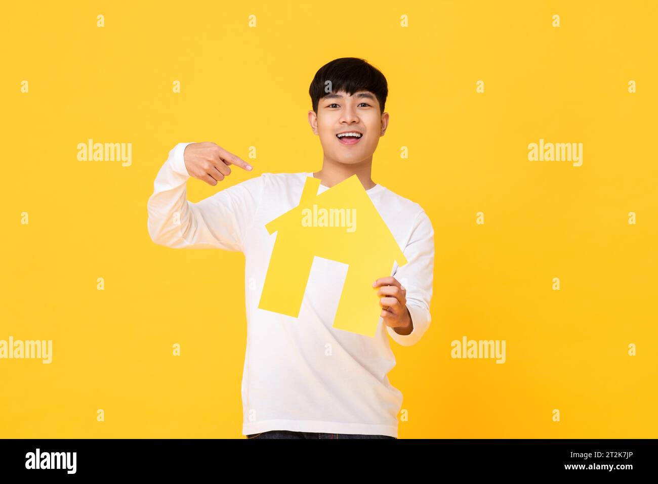 Souriant bel homme asiatique tenant et pointant la main à la maison modèle sur fond isolé de studio jaune Banque D'Images