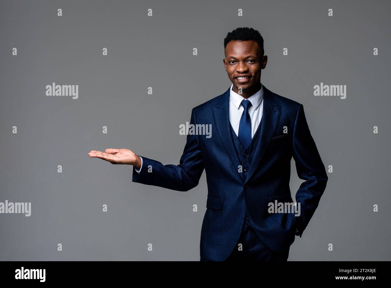 Beau jeune homme d'affaires africain portant costume d'affaires formel souriant et ouvrant la main dans le fond isolé de couleur gris studio Banque D'Images