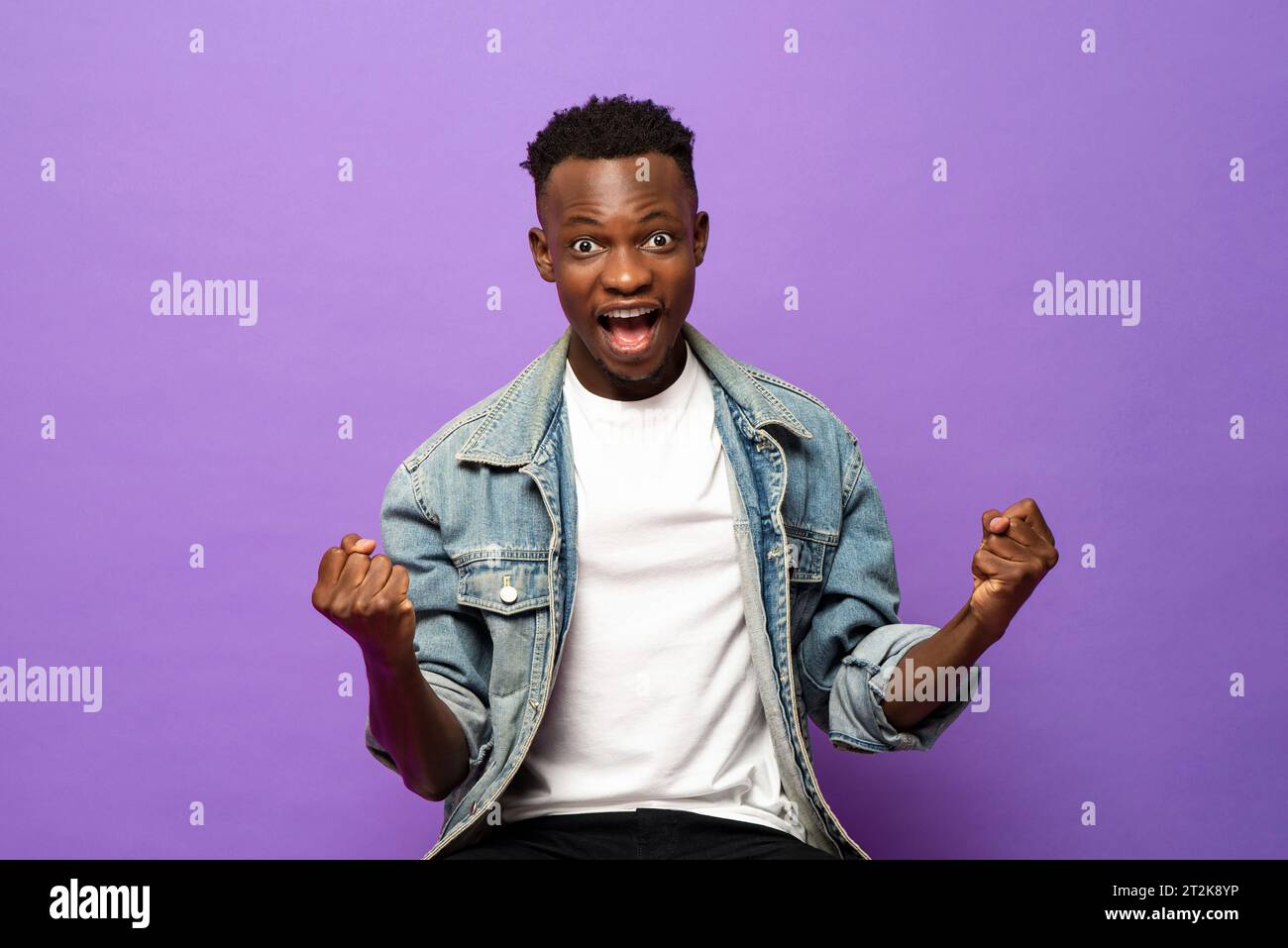 Gagnant extatique jeune homme africain serrant ses poings avec le visage excité dans le fond isolé de studio de couleur violette Banque D'Images