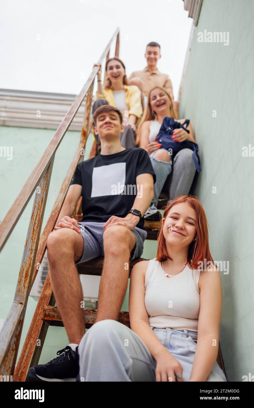 Des adolescents souriants drôles en vêtements décontractés sont assis sur une échelle métallique sur le toit d'un bâtiment. Les amis mignons s'amusent ensemble. Filles et garçons heureux Banque D'Images