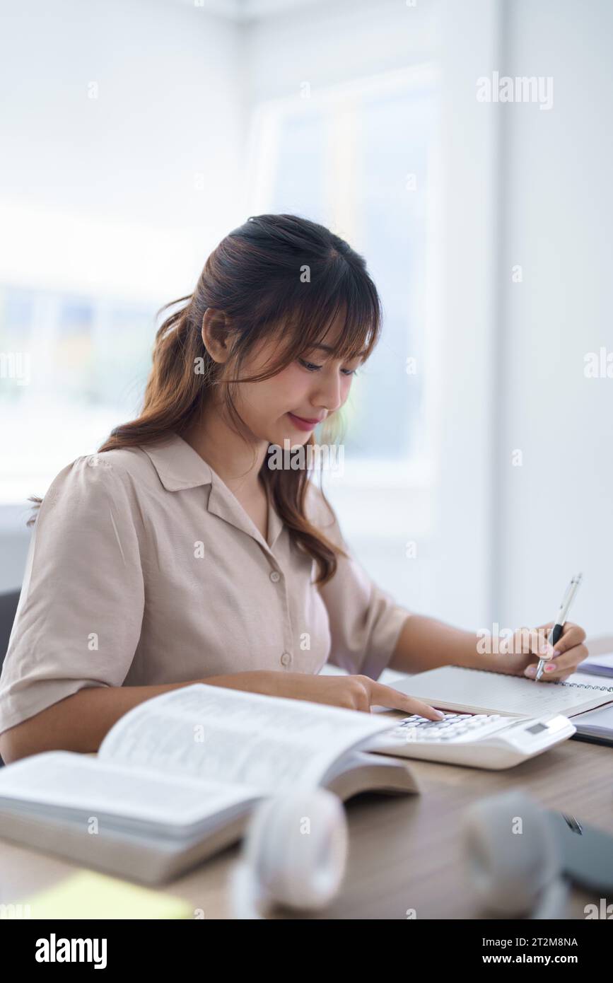 Jeune étudiant asiatique en vêtements décontractés utilisant la calculatrice pour faire des devoirs et écrire des notes dans un cahier après avoir étudié les connaissances de l'éducation Banque D'Images