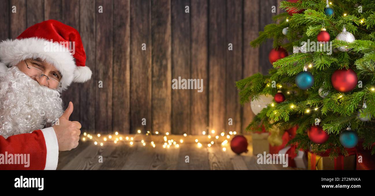 Le Père Noël en clin d'œil montre les pouces vers le haut et présente votre texte ou contenu sur un espace copie en bois sombre entre lui, un sapin de Noël et une lumière éclatante Banque D'Images