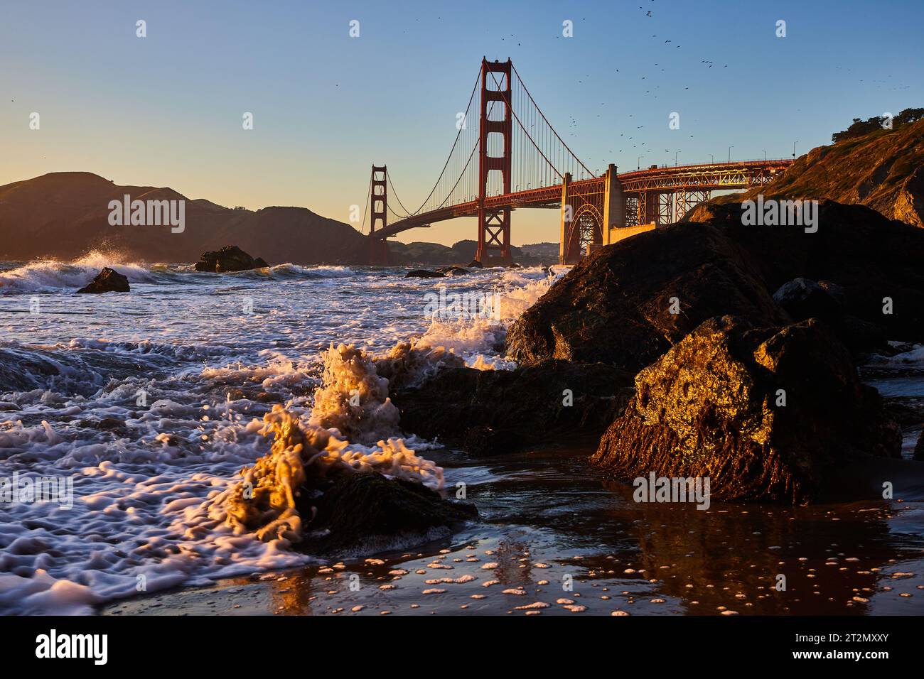 Golden Seafoam pulvérisant autour du rocher noir sur la rive avec Golden Gate Bridge à distance Banque D'Images