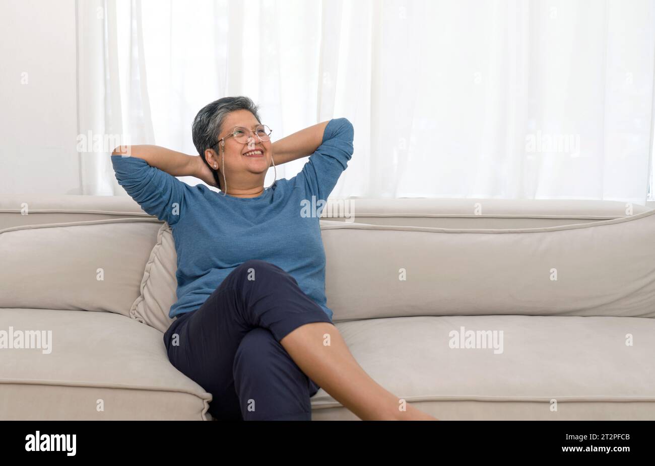 Une dame âgée sereine niche tranquillement sur son canapé en peluche, ourlée dans son salon élégamment éclairé, immergée dans ses pensées favorisant la tranquili Banque D'Images