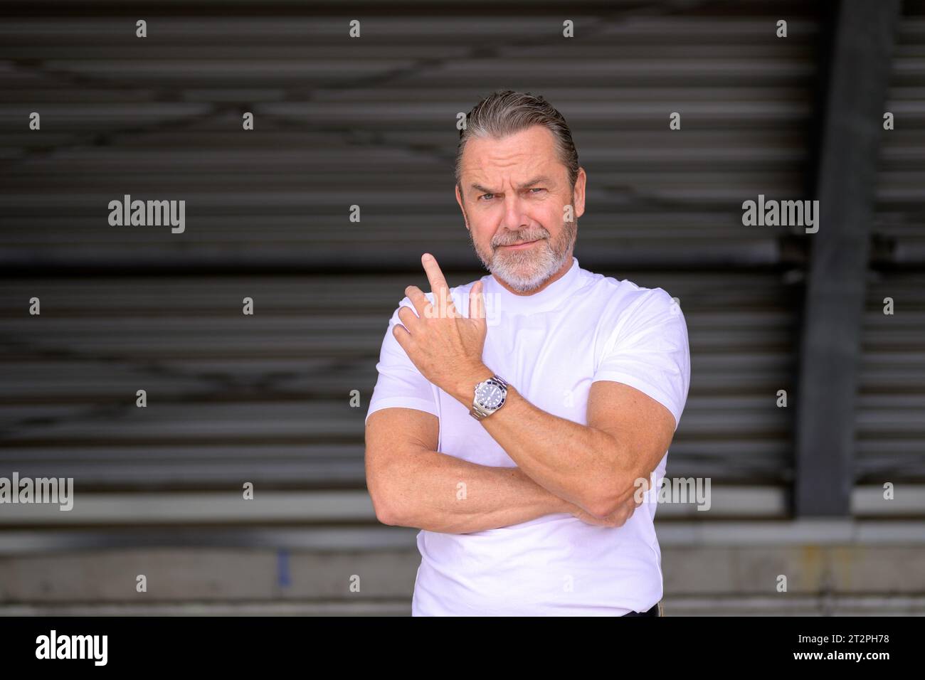 Attrayant homme aux cheveux gris avec un t-shirt blanc portant une montre de fantaisie et regardant de manière critique et demandant à la caméra, devant un fer ondulé W Banque D'Images