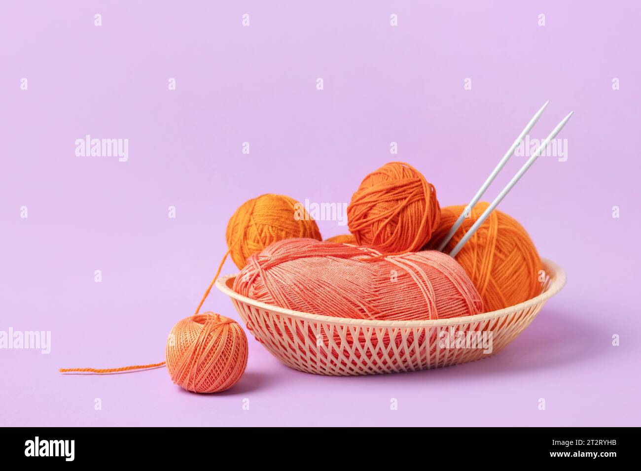 Boules à tricoter orange et rose dans un panier avec des aiguilles à tricoter sur fond lilas. Ensemble pour le passe-temps de tricoter des vêtements chauds ou des jouets. Besoin de concept Banque D'Images