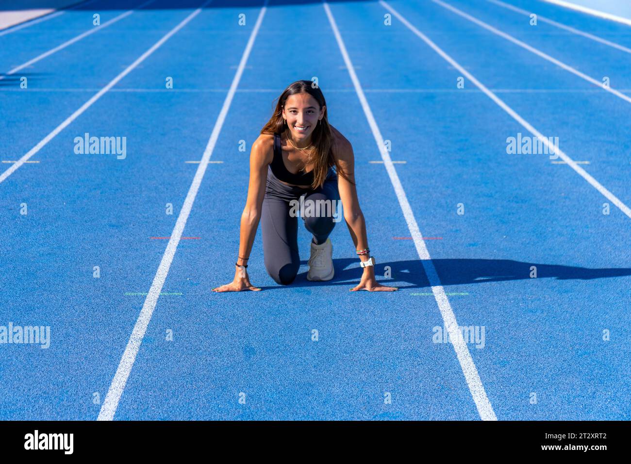 Belle jeune coureuse, bronzée avec de longs cheveux foncés, souriant sur une piste de course bleue regardant la caméra avec ses mains reposant sur le sol et la jambe Banque D'Images