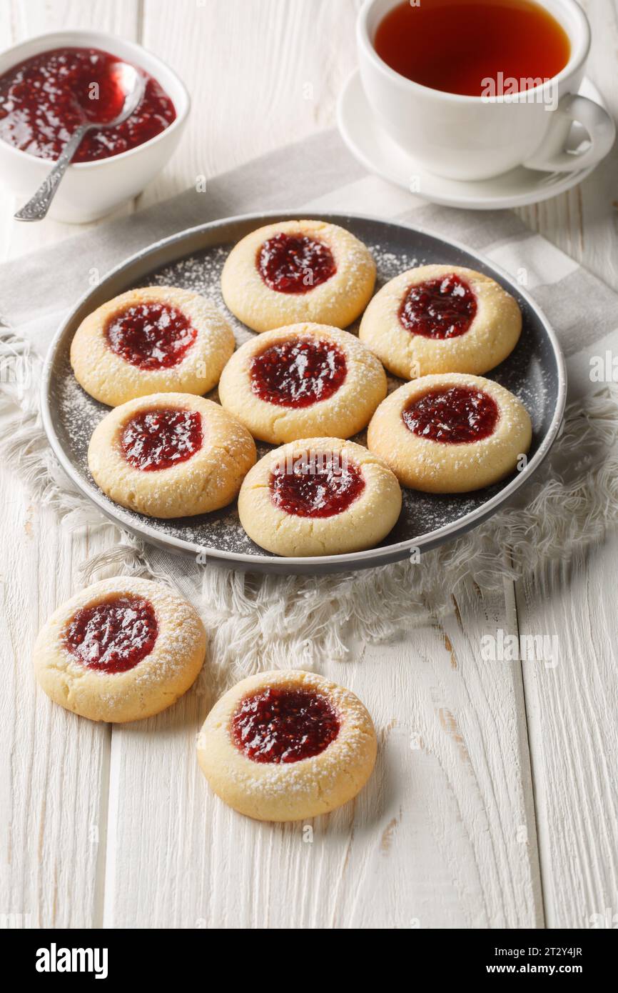 Biscuits de Noël à empreinte de pouce remplis de confiture de framboise closeup sur l'assiette servis avec du thé sur la table en bois blanc. Vertical Banque D'Images