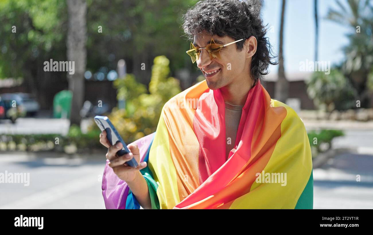 Jeune homme hispanique portant drapeau arc-en-ciel à l'aide d'un smartphone dans la rue Banque D'Images