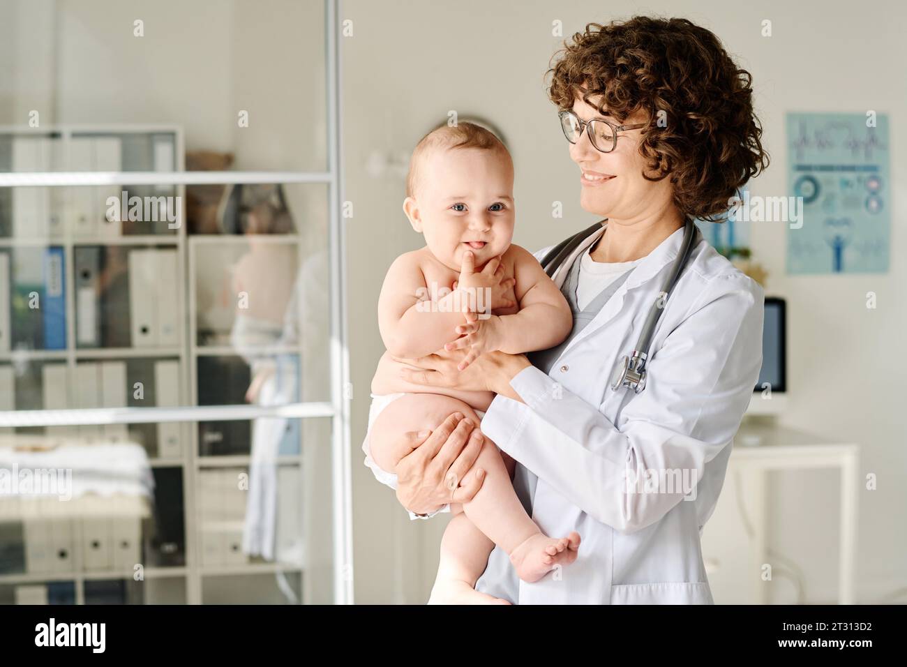Médecin souriant en manteau blanc tenant bébé sur les bras tout en se tenant au bureau Banque D'Images