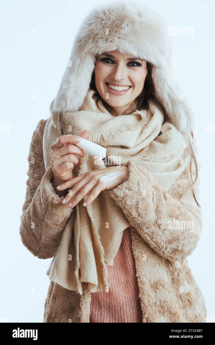 souriante femme moderne de 40 ans en manteau d'hiver et chapeau de fourrure isolé sur blanc avec crème pour les mains. Banque D'Images
