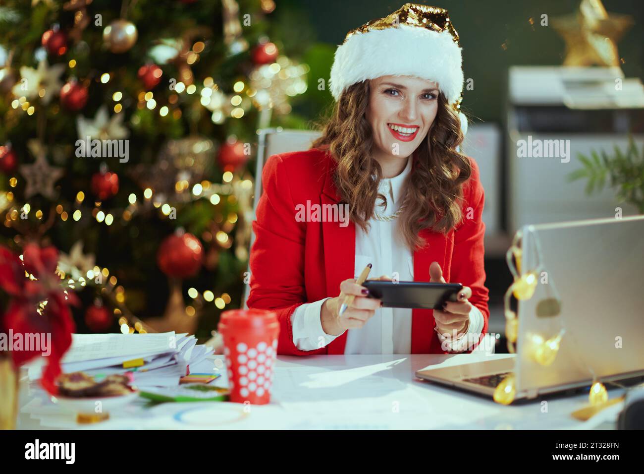 Noël. Portrait de heureux élégant dans la veste rouge avec ordinateur portable à l'aide de l'application smartphone et de travailler dans le chapeau de santa dans le bureau vert moderne avec Christma Banque D'Images
