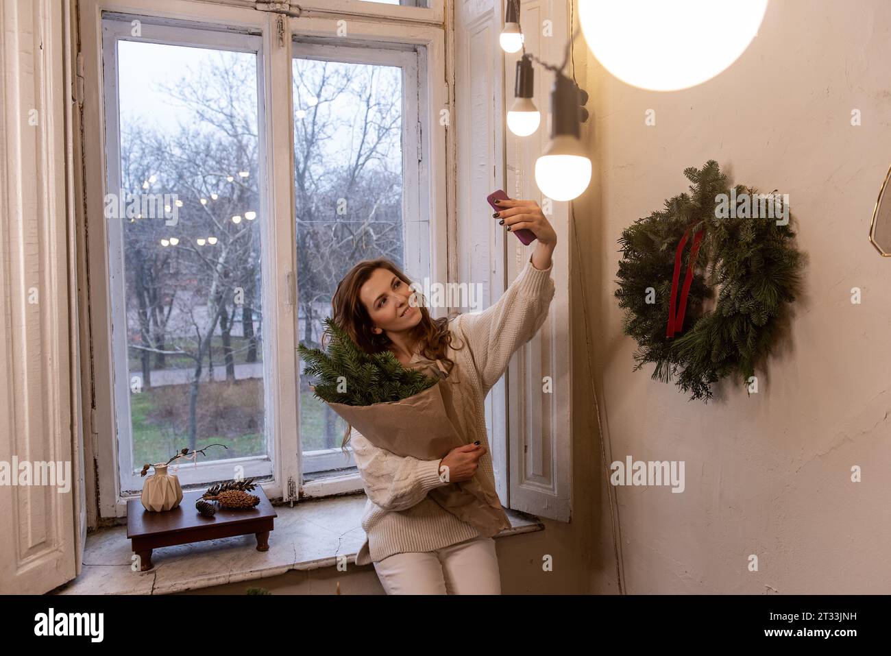 Femme élégante en pull blanc prend selfie sur smartphone, tient dans les mains bouquet de Noël en papier artisanal près de la fenêtre. Jeune fille millénaire ayant f Banque D'Images
