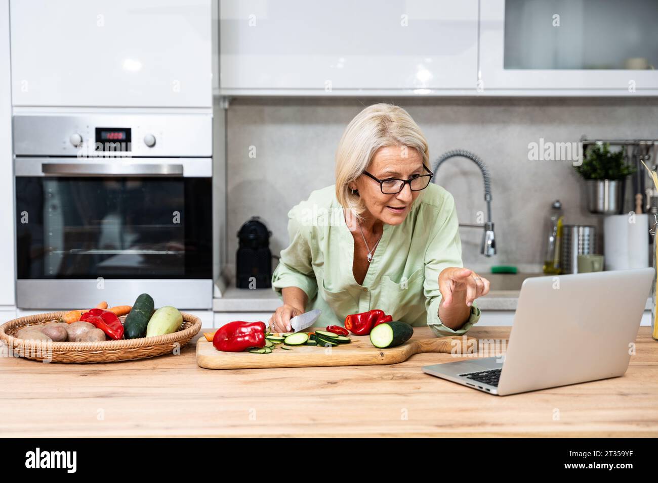 Femme senior à la recherche de recettes de cuisine en ligne sur son ordinateur portable. La femme retraitée cuisinent des légumes comme nourriture végétalienne debout dans l'ingrédient de coupe de cuisine Banque D'Images