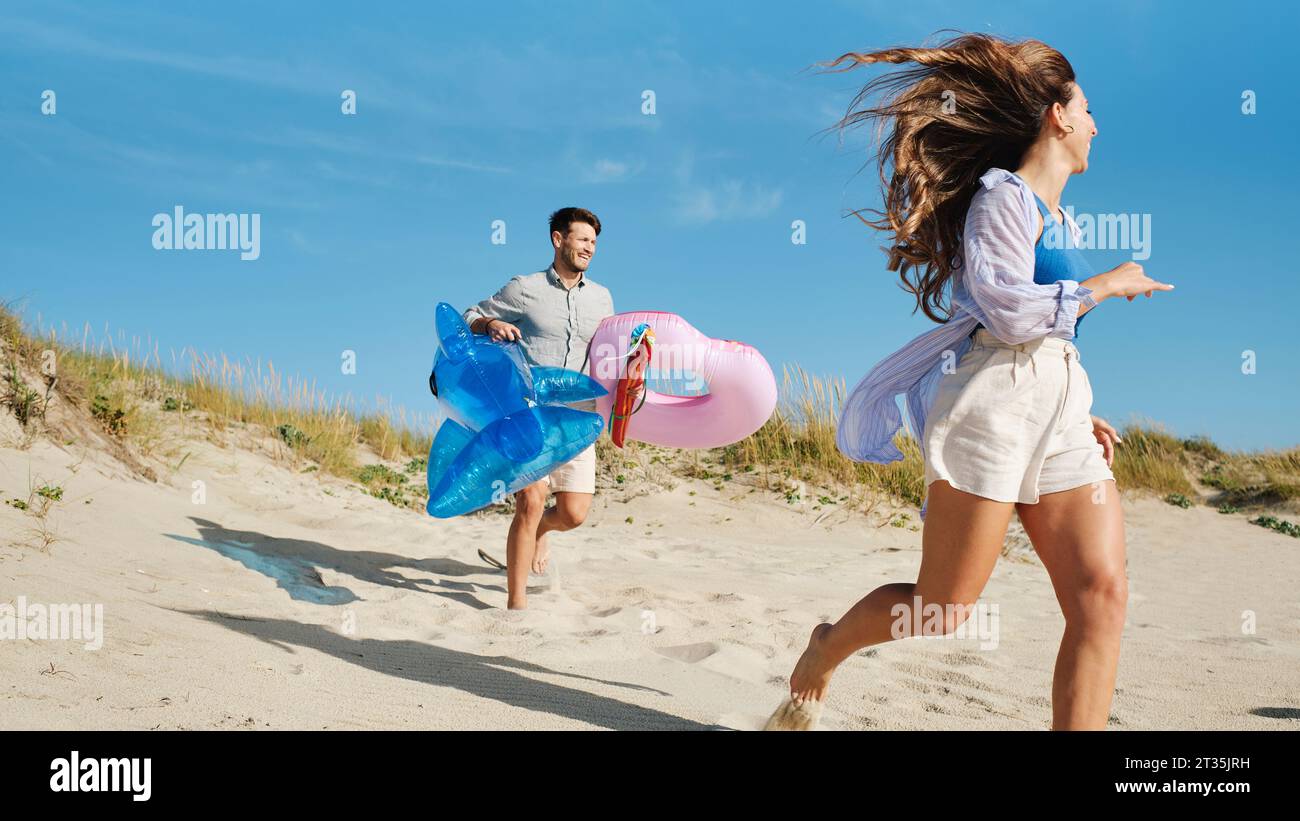 Femme heureuse courant avec l'homme tenant l'anneau de natation gonflable et le requin jouet Banque D'Images