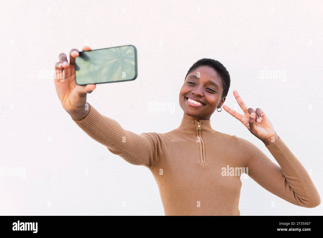 Femme heureuse montrant geste de signe de paix et prenant selfie devant le mur Banque D'Images